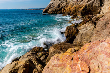 Fototapeta na wymiar The Mediterranean Sea waves reaching the big rocks on the shore with the sunlight reflecting in the turquoise water on a sunny day (Nice, France)