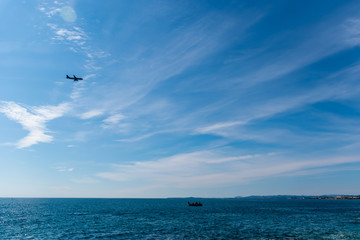 Panoramic wide angle shot of the Mediterranean Sea with the sunlight reflecting in the turquoise water, an airplane flying above it, a boat sailing, and the peninsula in the background (Côte d'Azur)