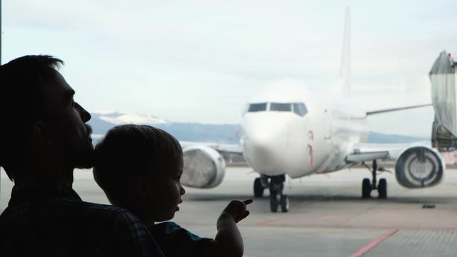 Silhouette of father and son looking and pointing by fingers at airplane through the window of   airport terminal. Family journey, travel with child lifestyle.
