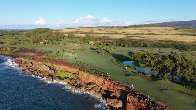 Low elevation aerial video of the Makawehi Bluff in the southeast coast of Kauai with lava cliffs, green golf courses, blue ocean and a mountain range. Poipu, Koloa, Kauai