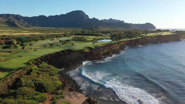 Low elevation aerial video of the Makawehi Bluff in the southeast coast of Kauai with lava cliffs, green golf courses, blue ocean and a mountain range, backlit. Poipu, Koloa, Kauai