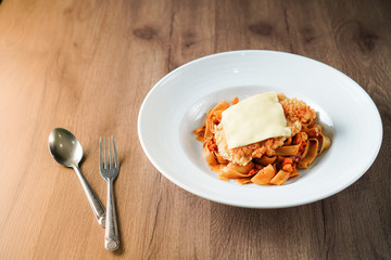 Pasta, Italian food, with tomato sauce topping with deep fried crispy pork cutlet and a sheet of cheese in white dish place on dark brown wooden table and stainless steel utensil near it