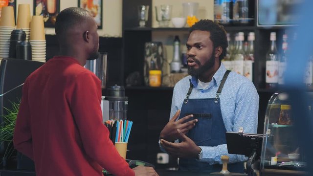 black bearded man in apron stands behind bar counter and talks with client from native country in cafe
