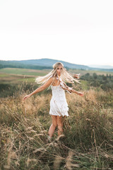 Woman fashion model portrait outdoors in the summer field, sunset. Boho style young woman in white dress and with headdress made of feathers
