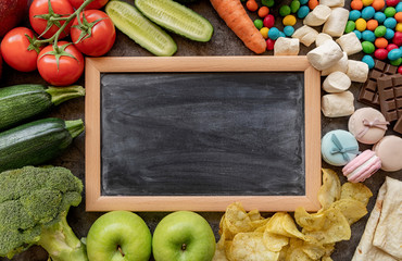 Fruit and vegetables vs sweets and potato fries around the blank chalk board top view flat lay