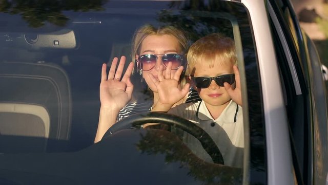 Mom and son in sunglasses sitting together at the wheel of the car, the child is played sitting on the hands of the mother in the car at sunset. Mother and son together waving their hands. Slow motion