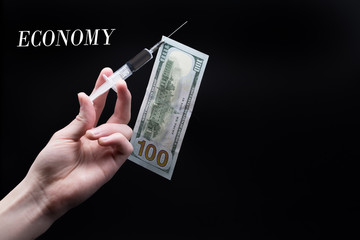 Hand holds a medical syringe. A dollar bill hangs on a needle. The inscription "economy". The dependence of the economy on oil prices.