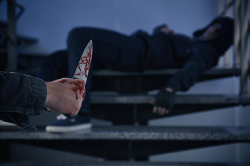 Man with bloody knife and unconscious person on stairs indoors, space for text. Dangerous criminal