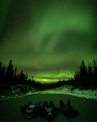 Three people laying watching Aurora Borealis Northern Lights in Yukon Territory Canada near Whitehorse with bright green bands covering the sky with snow on ground.