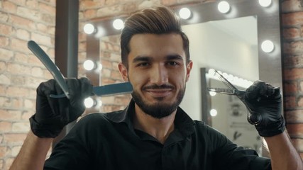 Smiling hairdresser snapping scissors front camera in fashion barbershop. Portrait winking barber with straight razor and scissors posing to camera in hair salon