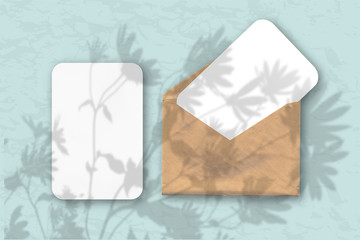 An envelope with 2 sheets of white textured paper on a pastel blue wall background. Mockup with an overlay of plant shadows. Natural light casts shadows from flowers and leaves of daisies