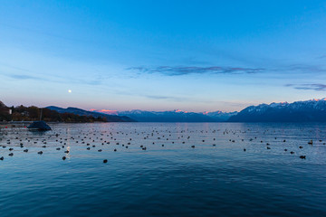 Obraz na płótnie Canvas Stunning panorama view of Lake Leman (Lake Geneva) at dusk in autumn in Lausanne, with beautiful snow covered French Alps mountain range and blue sky moon in background, Canton of Vaud, Switzerland