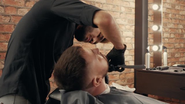Barber shaving neck to bearded man with straight razor in barbershop. Hairdresser using straight razor for beard shave in beauty salon. Handsome man getting beard care in male salon