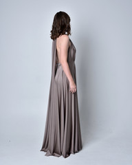  Portrait of a pretty brunette girl wearing a long silver evening gown, full length standing pose against a studio background.