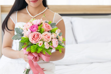 Obraz na płótnie Canvas An Asian bride in a wedding dress is sitting smiling brightly on the bed in her hand holding a beautiful bouquet of flowers.