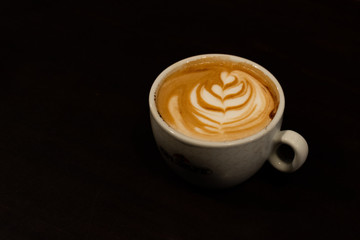 A cup of cappuccino with a flower in the milk foam on black background - latte art coffee shop concept