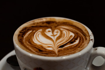 A cup of cappuccino with a flower in the milk foam on black background - latte art coffee shop concept