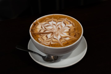 A cup of cappuccino with a flower in the milk foam on black background - latte art coffee shop concept
