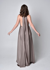 Fototapeta premium Portrait of a pretty brunette girl wearing a long silver evening gown, full length standing pose against a studio background.