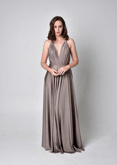 Fototapeta premium Portrait of a pretty brunette girl wearing a long silver evening gown, full length standing pose against a studio background.