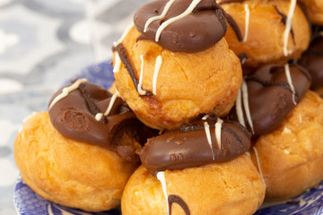 Profiteroles, coated in chocolate, stacked on a pattermed blue and white plate.  On a tile background