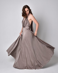  Portrait of a pretty brunette girl wearing a long silver evening gown, full length standing pose against a studio background.