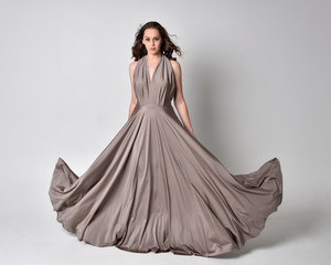  Portrait of a pretty brunette girl wearing a long silver evening gown, full length standing pose against a studio background. © faestock