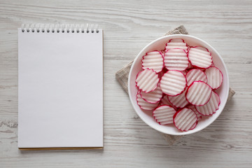 Radish Chips Slices in a white bowl, blank notepad on a white wooden surface, top view. Flat lay, overhead, from above. Copy space.