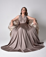 Portrait of a pretty brunette girl wearing a long silver evening gown, full length seated pose against a studio background.