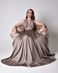 Fototapeta premium Portrait of a pretty brunette girl wearing a long silver evening gown, full length seated pose against a studio background.