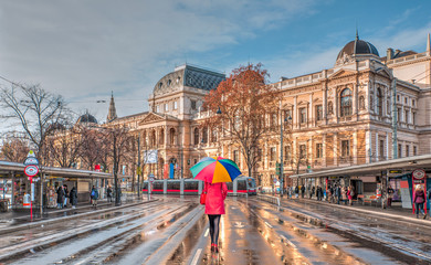 Woman in red clothes with multicolored umbrella - Tram moving on a street  - View of the University of Vienna (Universitat Wien) - Austria - obrazy, fototapety, plakaty