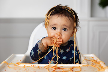 Funny baby in high chair eating spaghetti with his hands