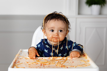 Funny baby with messy face and spaghetti hanging from mouth