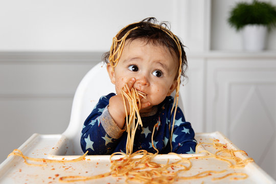 Cute baby in high chair eating spaghetti with his hands