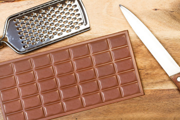 Tableta de chocolate con cuchillo y rallador sobre una tabla de madera rústica. Vista superior