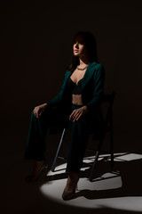Studio photo of pretty brunette woman in twilight sitting on black chair. A ray of light hits her face.