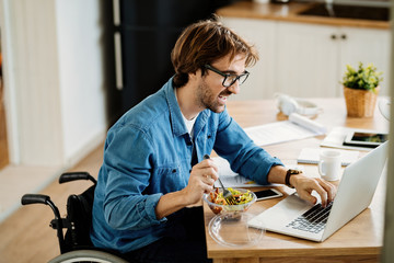 Happy businessman in wheelchair eating salad while working on laptop at home.