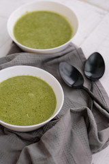Spring detox broccoli green cream soup in white plates, bowl with crackers and stylish black cutlery on grey background. 