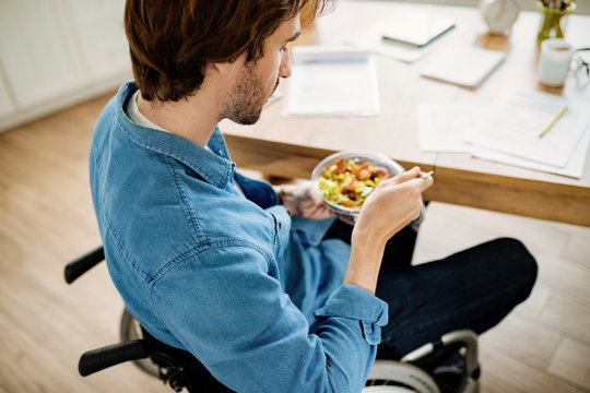 Young Man In Wheelchair Eating While Working At Home.