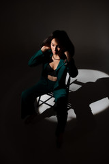 Studio photo of pretty brunette woman in twilight sitting on black chair. A ray of light hits her face.