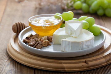 White, round cheeses on wooden background with honey, nuts and grapes. Dark food photo.