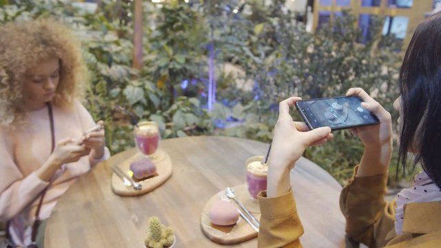 Two young female friends taking pictures of fruity cocktails and cakes with smartphones while sitting together at cafe table beside green plants
