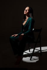 Fototapeta premium Studio photo of pretty brunette woman in twilight sitting on black chair. A ray of light hits her face.