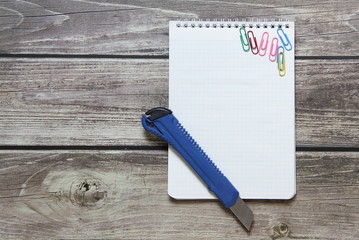 Notepad with a blank white sheet in a checker paper and with stationery knife lies on the background of wooden boards.