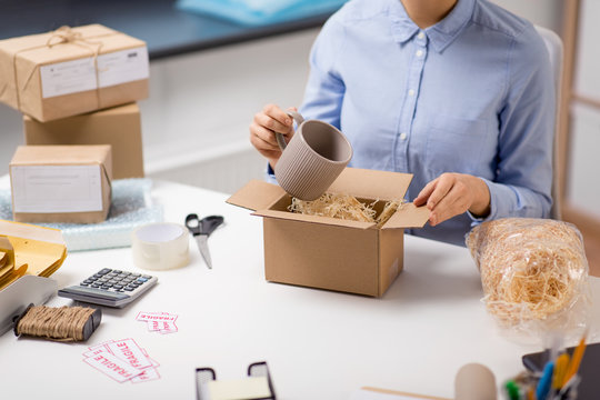 delivery, mail service, people and shipment concept - close up of woman packing mug to parcel box with straw filler at post office