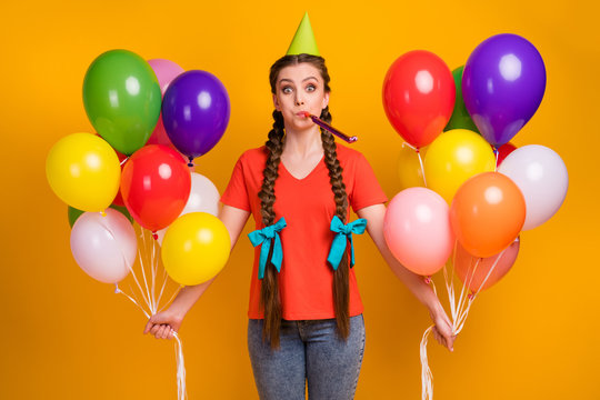 Photo of attractive funny lady hold many air balloons hands birthday party best friend blow noisemaker wear paper cone cap casual red t-shirt jeans isolated vibrant yellow color background