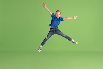 Jumping high. Happy boy playing and having fun on green studio background. Caucasian kid in bright cloth looks playful, laughting, smiling. Concept of education, childhood, emotions, facial expression