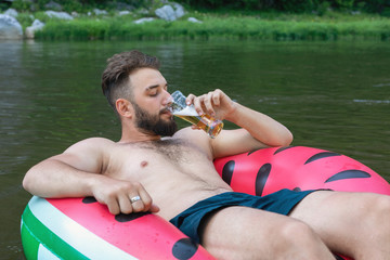 Portrait of bearded hipster guy floating on inflatable circle with glass of beer in his hand....