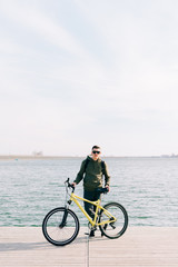 Obraz premium a young man in khaki sportswear and glasses stands on a pontoon near the lake and holds a yellow Bicycle near him on a Sunny spring day