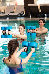 Fototapeta premium Selective focus of smiling man and woman looking at trainer with barbells in swimming pool